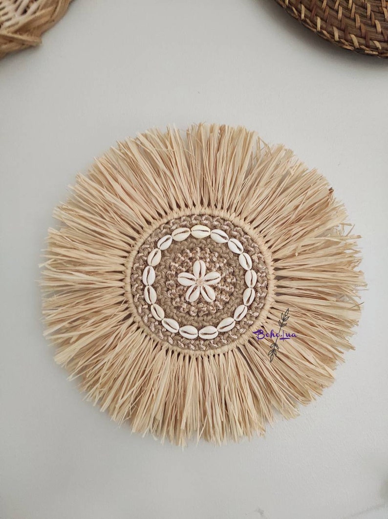 May include: A round, natural-toned woven wall hanging with a central design. The center features a star-shaped design made of shells, surrounded by a ring of shells and a woven border. The outer edge has a fringe.