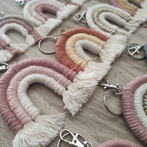 Puede incluir: Una colección de llaveros de arcoíris de macramé hechos a mano en varios colores pastel, incluyendo rosa, beige y dorado. Cada llavero tiene un cierre y un anillo de metal. Los llaveros están colocados sobre una superficie de madera.