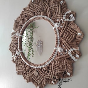 Könnte beinhalten: Runder Makramee-Spiegel mit einem braunen und weißen Webmuster. Der Rahmen hat aufwendige Knotenarbeit, weiße Akzente und kleine Muscheln. Dieser dekorative Wandbehang hat einen Boho-Stil.