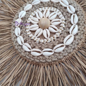 May include: A round, woven wall hanging with a natural, earthy aesthetic. The center features a circle of white seashells, a wooden sphere, and gold-colored beads. The outer edge has a fringe of dried palm leaves.