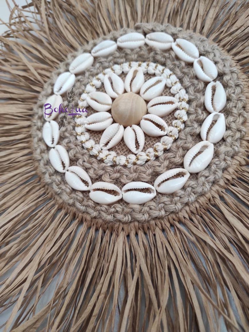 May include: A round, woven wall hanging with a natural, earthy aesthetic. The center features a circle of white seashells, a wooden sphere, and gold-colored beads. The outer edge has a fringe of dried palm leaves.
