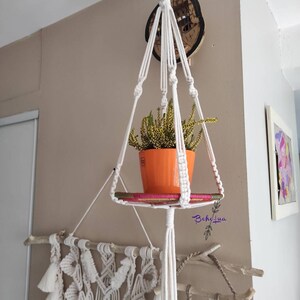 Peut inclure: Suspension murale en macramé avec un pot orange contenant une plante à fleurs jaunes. L'étagère est soutenue par des cordons en macramé blanc et une cheville en bois. Le design comprend une tenture murale décorative en macramé.