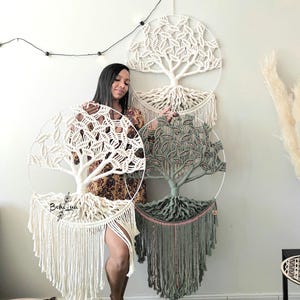 May include: Macramé wall hangings with tree designs. The circular frames feature intricate knotting in cream and green, with fringed bottoms. The wall art is displayed against a neutral wall.