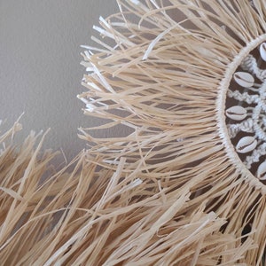 May include: Close-up of a natural fiber wall hanging. The circular design features a woven center adorned with seashells, surrounded by a fringe of long, beige strands. The texture and color suggest a coastal or bohemian aesthetic.
