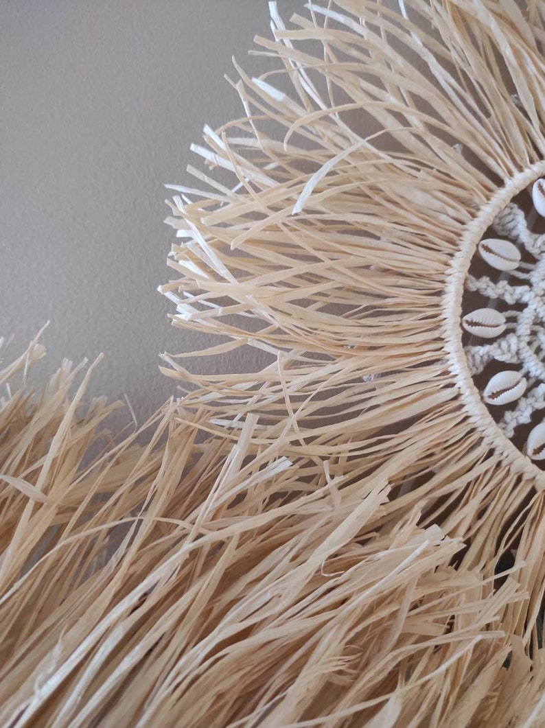 May include: Close-up of a natural fiber wall hanging. The circular design features a woven center adorned with seashells, surrounded by a fringe of long, beige strands. The texture and color suggest a coastal or bohemian aesthetic.