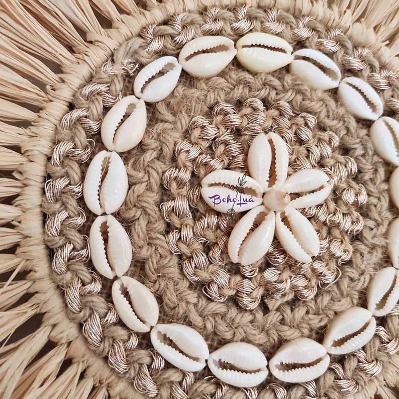 May include: A round, woven coaster with a natural, beige color. The coaster is decorated with white seashells arranged in a circular pattern around the edge and a flower shape in the center. The text "BohoLua" is visible.