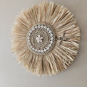 May include: A round, natural fiber wall hanging with a central woven design. The center is adorned with small seashells and a flower-shaped accent. The outer edge features long, radiating strands of dried grass, creating a sunburst effect. The wall decor has the text "BohLua".