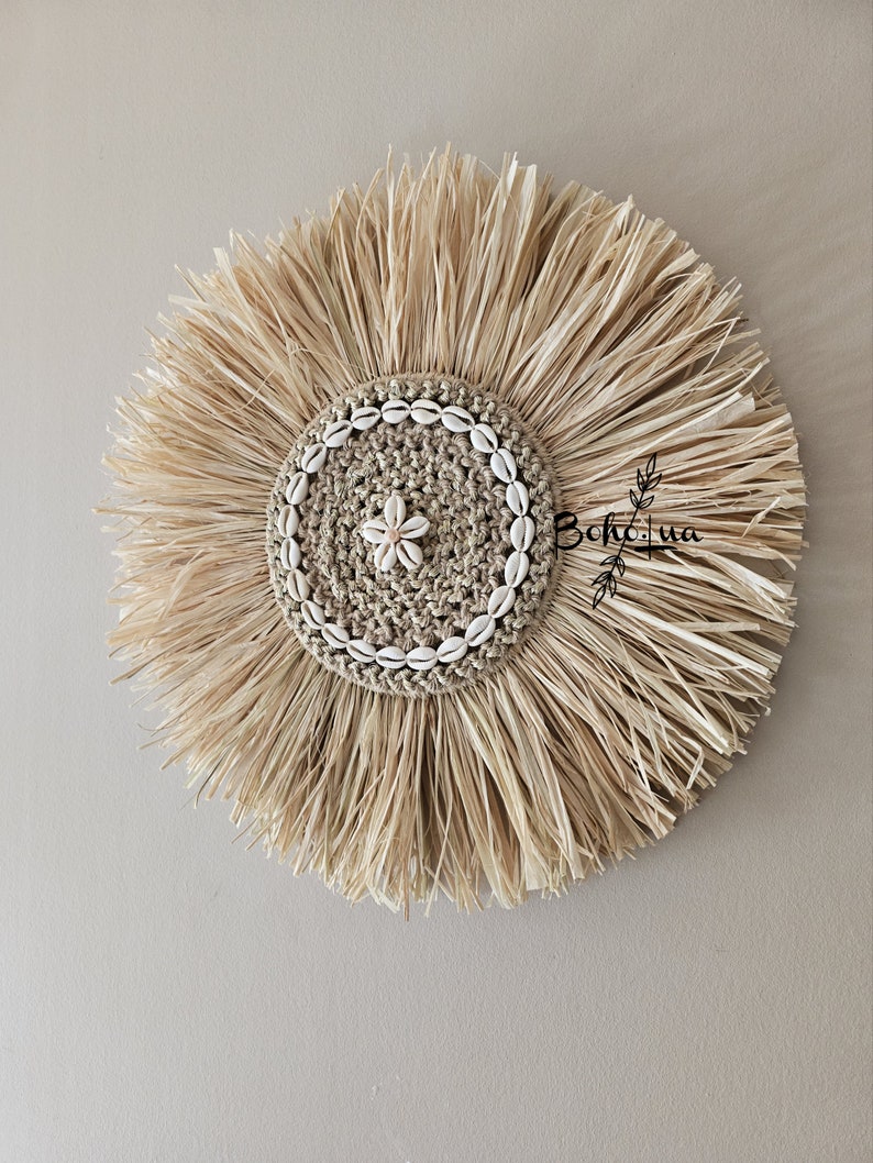 May include: A round, natural fiber wall hanging with a central woven design. The center is adorned with small seashells and a flower-shaped accent. The outer edge features long, radiating strands of dried grass, creating a sunburst effect. The wall decor has the text "BohLua".