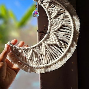 Op de afbeelding: Crèmekleurige macramé halve maan wandhanger met een sterontwerp en franjes. Een klein, iriserend kristallen prisma hangt onder de maan. De tekst "Bohe_ma" is zichtbaar.