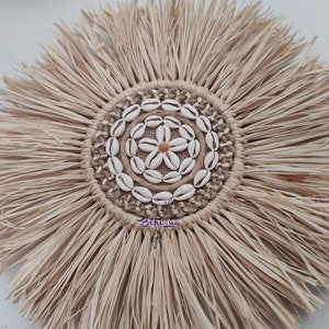 May include: A round, natural-toned wall hanging with a central design of seashells arranged in a flower pattern. The center is surrounded by a ring of woven jute and gold thread. The outer edge features long, fringed raffia.