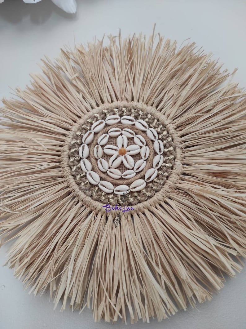 May include: A round, natural-toned wall hanging with a central design of seashells arranged in a flower pattern. The center is surrounded by a ring of woven jute and gold thread. The outer edge features long, fringed raffia.