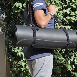Peut inclure: Un tapis de yoga noir enroulé, maintenu par une sangle en macramé noire. La sangle est ornée d'anneaux en bois et d'un motif tressé. Le tapis est porté à l'épaule, facilitant le transport pour les activités sportives.