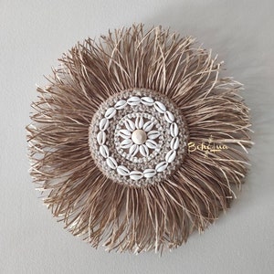May include: A round, woven wall hanging with a central design of seashells and a natural fiber fringe. The center features a circular pattern of small white seashells around a larger shell. The fringe is made of long, thin strands of a light brown material.