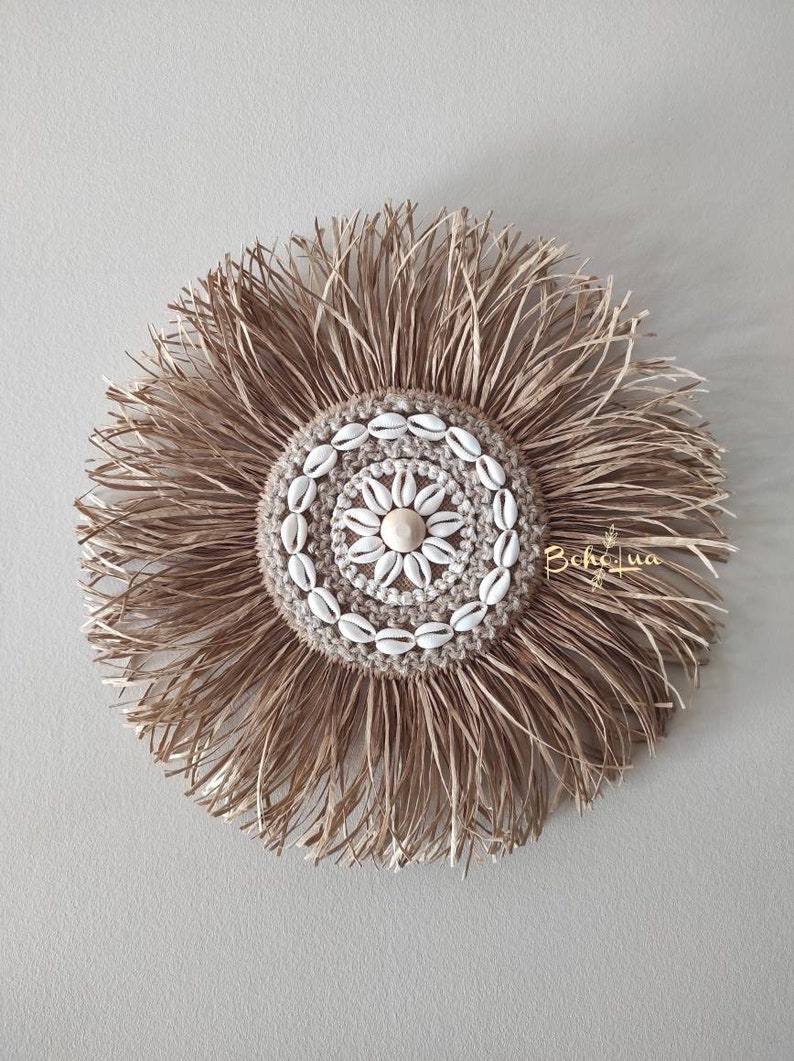 May include: A round, woven wall hanging with a central design of seashells and a natural fiber fringe. The center features a circular pattern of small white seashells around a larger shell. The fringe is made of long, thin strands of a light brown material.