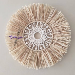 May include: Circular wall decor featuring a natural, beige woven design with a central white macrame pattern. The macrame is adorned with seashells. The outer edge has a fringe of natural fibers. The decor has a boho style.