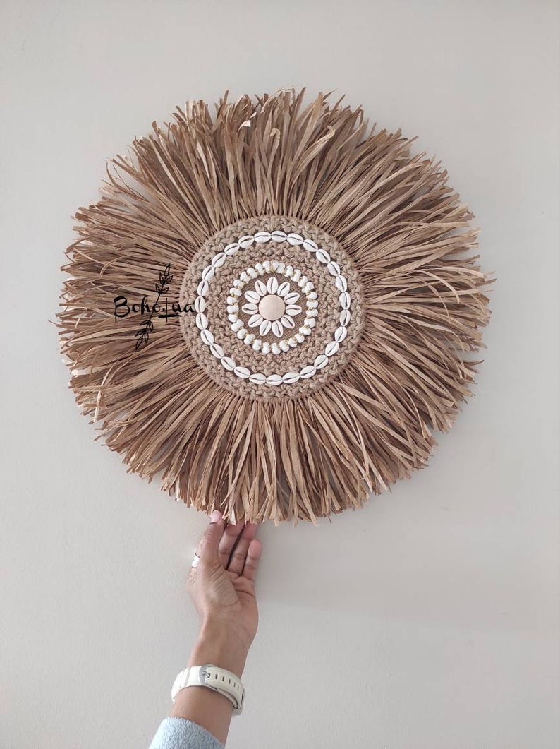 May include: A round, natural fiber wall hanging with a central design of seashells and a woven jute center. The outer edge features long, fringed fibers. The piece is held by a person's hand, showcasing its size and texture. The wall decor has a boho style.