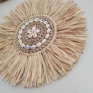 May include: A round, natural fiber wall hanging with a central design of seashells and a flower-shaped shell. The outer edge features a fringed, sunburst design. The word "Bohohua" is written on the hanging.