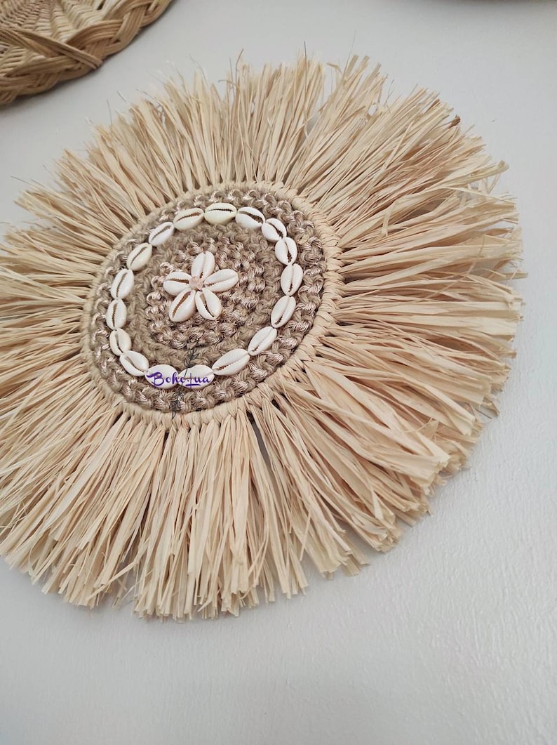 May include: A round, natural fiber wall hanging with a central design of seashells and a flower-shaped shell. The outer edge features a fringed, sunburst design. The word "Bohohua" is written on the hanging.