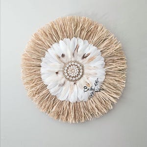 Puede incluir: Decoración de pared redonda con un diseño central de plumas blancas y un centro adornado con conchas. Las plumas están rodeadas por un anillo de paja de color natural. La palabra "Boho Lua" está escrita en escritura negra en el lado derecho.
