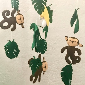 May include: A baby mobile with a jungle theme. It features three brown felt monkeys, green felt leaves, and a yellow felt banana.