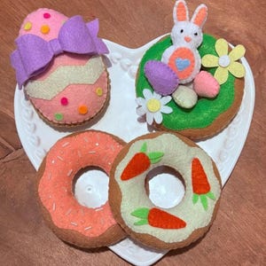 Op de afbeelding: Vier vilten donuts met een paasthema op een wit hartvormig bord. Eén donut is versierd met een roze en wit ei, een paarse strik en kleurrijke sprinkels. Een andere donut is versierd met groene glazuur, een wit konijn en paarse en roze eieren. De andere twee donuts zijn versierd met witte glazuur en oranje wortels.