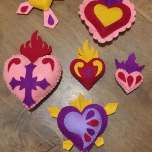 Può includere: Sei ornamenti a forma di cuore in feltro con diversi design. I cuori sono rosa, rossi e viola con accenti gialli, rossi e viola. Alcuni cuori hanno fiamme, croci e corone.