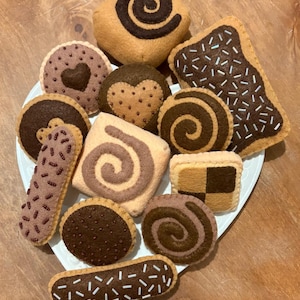 May include: A collection of felt cookies in various shapes and colors, including swirls, hearts, and sprinkles. The cookies are arranged on a white plate.