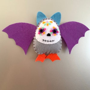 May include: A felt bat with purple wings, blue ears, and orange feet. The bat's face is white with colorful floral accents and a stitched black mouth. The wings are stitched with purple thread.