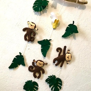 May include: A brown and green felt mobile with three monkeys, a banana, and leaves hanging from a driftwood branch.