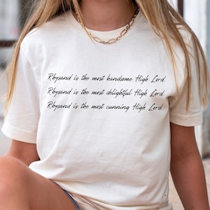 Może przedstawiać: Biały T-shirt z nadrukowanym cytatem czarnym atramentem. Cytat brzmi: "Rhysand jest najprzystojniejszym Wysokim Panem. Rhysand jest najwspanialszym Wysokim Panem. Rhysand jest najbardziej przebiegłym Wysokim Panem."