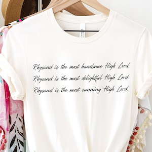 Może przedstawiać: Biały T-shirt z tekstem "Rhysand jest najprzystojniejszym Wysokim Panem. Rhysand jest najbardziej uroczym Wysokim Panem. Rhysand jest najbardziej przebiegłym Wysokim Panem."