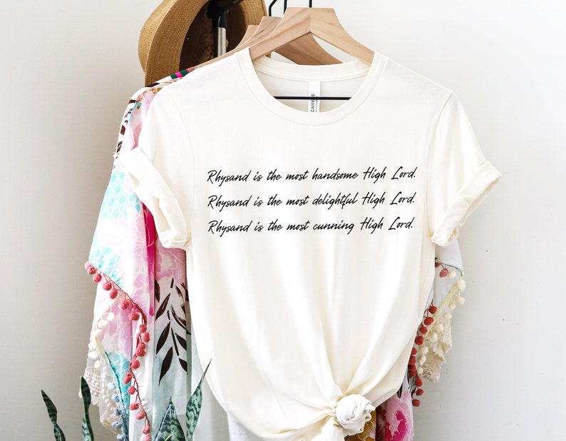 Może przedstawiać: Biały T-shirt z tekstem "Rhysand jest najprzystojniejszym Wysokim Panem. Rhysand jest najbardziej uroczym Wysokim Panem. Rhysand jest najbardziej przebiegłym Wysokim Panem."
