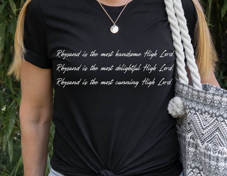 Może przedstawiać: Czarny T-shirt z białym tekstem, kt&oacute;ry brzmi: "Rhysand jest najprzystojniejszym Wysokim Panem. Rhysand jest najbardziej uroczym Wysokim Panem. Rhysand jest najbardziej przebiegłym Wysokim Panem."