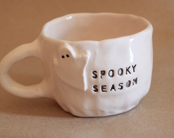 May include: A white ceramic mug with an irregular, hand-formed shape and a curved handle. The mug features a small, raised ghost figure with two black dots for eyes and the text "SPOOKY SEASON" stamped in black letters on the side.