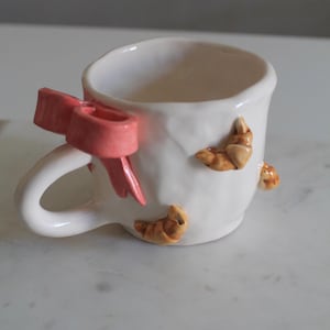 May include: A white ceramic mug with a pink bow on the handle and croissant-shaped decorations. The mug features a curved handle and a smooth, glossy finish. The bow is a soft, pastel pink, adding a playful touch to the design.