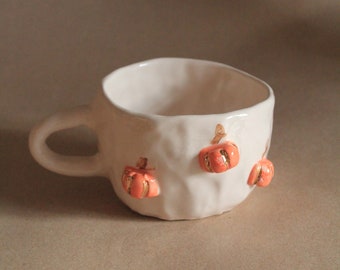 May include: A white ceramic mug with an irregular, textured surface and a single handle. The exterior is decorated with small, raised orange pumpkin motifs featuring gold-colored accents on the ridges of each pumpkin.