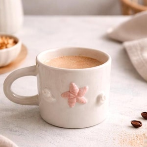 Könnte beinhalten: Ein weißer Keramikbecher mit einem abgerundeten Henkel, der auf der Vorderseite ein erhabenes, strukturiertes rosa Seestern-Emblem aufweist. Der Becher ist mit einer hellbraunen Flüssigkeit gefüllt und die Oberfläche ist glatt mit dezenten Details.
