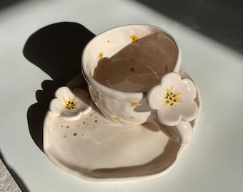 May include: A white ceramic teacup and saucer set with a floral design. The cup and saucer are decorated with small yellow flowers and a larger white flower with yellow accents on the handle.