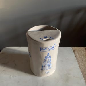Può includere: Una tasca da parete in ceramica bianca con illustrazioni blu disegnate a mano. Il design include un paesaggio urbano, cuori e le parole "ONE WAY" e "NY". La tasca ha una parte superiore curva e un retro piatto per appenderla.