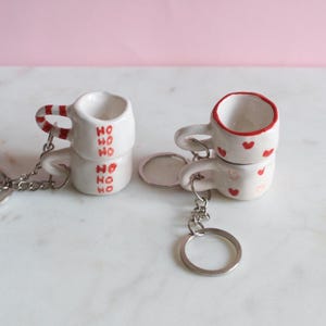 May include: Two stacked white ceramic mug keychains. One has red candy cane stripes on the handle and "HO HO HO" in red. The other has red heart accents and a red rim. Both have silver key rings.
