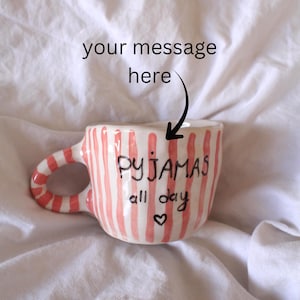 Puede incluir: Una taza de cerámica con acabado brillante que presenta rayas verticales de color rosa coral y una inscripción negra pintada a mano que dice "PYJAMAS all day" con un pequeño corazón. La taza incluye un asa curva con rayas rosas a juego.