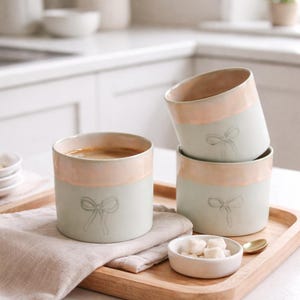 May include: Four ceramic mugs with a pale green base and a peach-coloured rim. Each mug has a bow design and are on a wooden tray with a small bowl of sugar cubes and a gold spoon. One mug is filled with coffee.