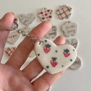 May include: A heart-shaped keychain with a white ceramic pendant featuring red strawberry illustrations and blue dots. The keychain is attached to a silver metal ring. Other ceramic pendants with various designs are visible in the background.
