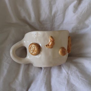 Può includere: Tazza in ceramica bianca con manico e dettagli marroni in rilievo di pasticcini, tra cui un croissant, un biscotto e un panino.