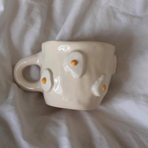 Könnte beinhalten: Eine weiße Keramiktasse mit Henkel, verziert mit erhabenen, 3D-Eiformungen mit gelben Eigelb. Die Tasse steht auf einer weißen Oberfläche.