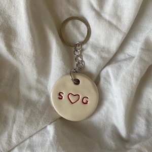 Könnte beinhalten: Weißer Keramik-Schlüsselanhänger mit einer silbernen Kette und einem roten Herzen mit den Buchstaben "SOG" auf beiden Seiten des Herzens.