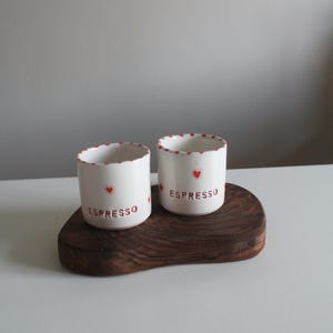 Könnte beinhalten: Zwei weiße Espresso-Tassen aus Keramik mit roten Herzakzenten und dem Aufdruck "ESPRESSO" auf einem dunklen Holztablett. Die Ränder der Tassen sind mit kleinen roten Herzen verziert.