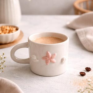 Puede incluir: Una taza de cerámica color crema con un diseño de flor rosa, llena de un líquido marrón claro. La taza tiene un asa curva y detalles en forma de concha. La taza está sobre una superficie blanca.