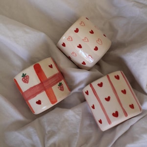 Puede incluir: Tres macetas cuadradas de cerámica con diseños pintados a mano. Una maceta presenta corazones rojos, otra rayas y corazones rojos, y la tercera fresas y un diseño de cruz roja. Las macetas son blancas con detalles rojos.