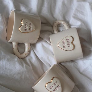 Könnte beinhalten: Drei weiße Keramikbecher mit goldenen Akzenten und rosa Beschriftung. Jeder Becher hat ein herzförmiges Design mit dem Text "BEST MOM" im Inneren.